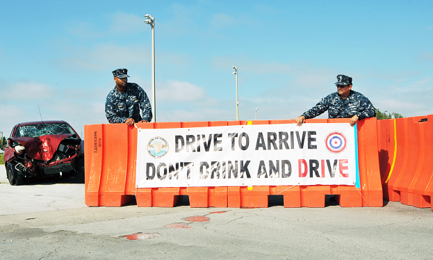 Zwei Polizeibeamte stehen neben einem beschädigten Auto mit einem Banner davor, unter einem klaren blauen Himmel mit Laternen und Bäumen im Hintergrund.