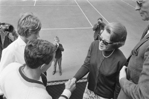 Eine Gruppe von Menschen auf einem Tennisplatz, mit einer Frau in einer Brille, die einem Mann in einem Anzug die Hand sch├Ąktelt, und Fotografen im Hintergrund, alles in Schwarz-Wei├č.