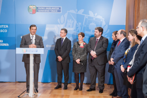 Eine Person in Anzug und Krawatte steht hinter einem Pult mit sichtbaren Abnutzungsspuren, flankiert von einer weiteren Person in schwarzem Anzug und Krawatte und einer Frau, mit einigen Personen im Hintergrund und einem Banner dahinter.