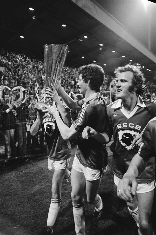 Schwarzes und weißes Bild von Männern, die auf einem Fußballfeld feiern und einen Pokal nach dem Gewinn des FA-Cup-Finals in die Höhe halten, mit sichtbaren Zuschauern hinter einem Zaun unter Stadionbeleuchtung.