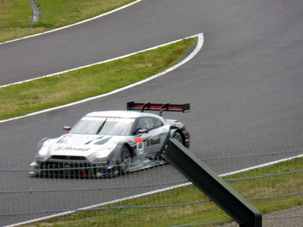 Ein Audi R8 LMS GT3 Rennauto ist auf der Nürburgring 24 Stunden Rennstrecke positioniert, umgeben von einem Metallzaun und grünem Gras, mit der Strecke, die sich vor ihm erstreckt.