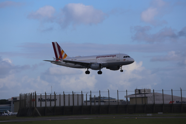Germanwings Airbus A320-200 startet am Frankfurter Flughafen mit Gras darunter, einem Zaun im Vordergrund, Gebäuden und Bäumen im Hintergrund und einer bewölkten Himmel.