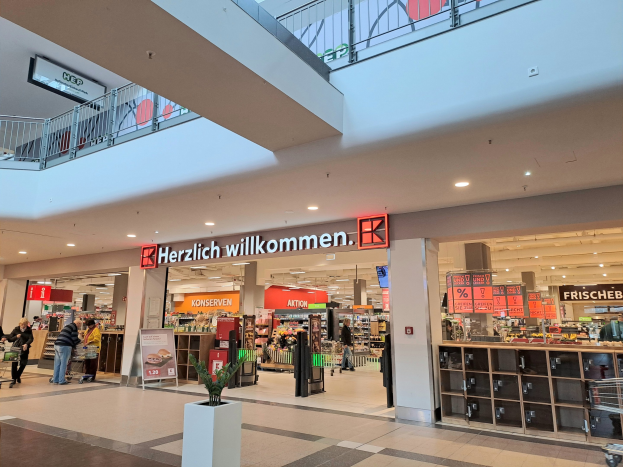 Innenraum eines Einkaufszentrums mit Menschen, Geschäften, beleuchteten Schildern einschließlich "Herzlich willkommen" (Deutsch für "Willkommen"), Geländern, Deckenleuchten und einem Topf mit Pflanze unten.