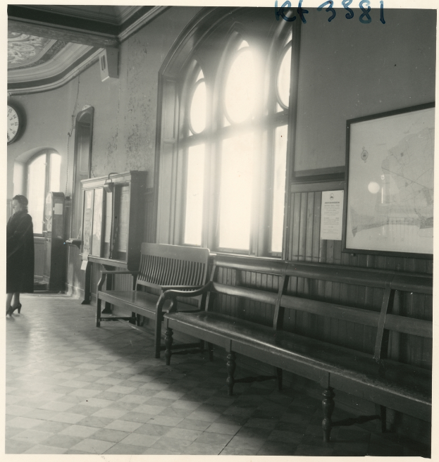 Ein Schwarz-Weiß-Foto einer Frau, die in einem Bahnhof steht, umgeben von Bänken, Schildern, Fenstern und einer Uhr an der Wand, mit Text oben auf dem Bild.
