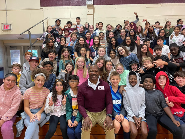 Gruppe von Schülern, die in einem Turnsaal für ein Foto posieren, einige sitzen auf der Treppe und andere stehen, eine Wand im Hintergrund, eine Tür und ein Ausgangsschild links.