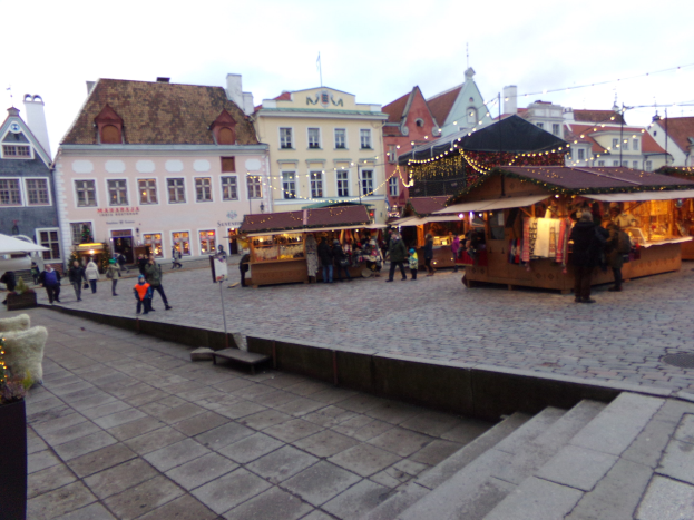 Ein belebter Weihnachtsmarkt in Tallinn, Estland, mit Menschen um geschmückte St├Ąnde, festlicher Beleuchtung, bew├Âlktem Himmel, Treppen und Topfpflanzen im Bereich.