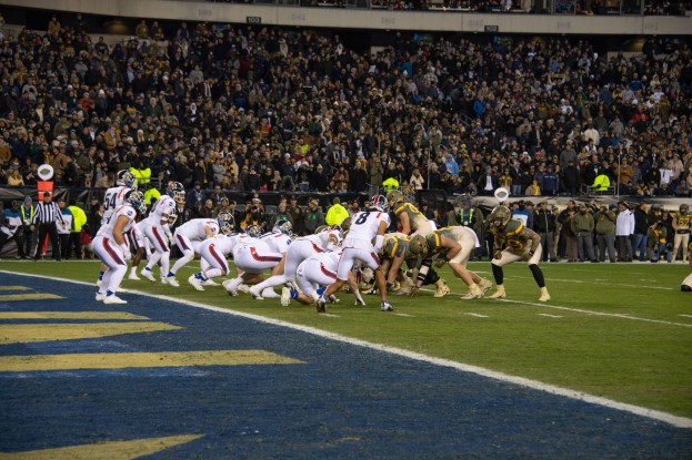 Ein Fußballspiel im Gange mit Spielern auf dem Rasen in Sportkleidung und Helmen, aus der Perspektive der Tribüne mit Zuschauern und Werbetafeln im Hintergrund.