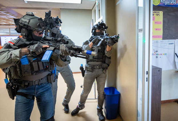 Gruppe von Polizisten in Uniform mit Helmen und Waffen in einem Raum mit einem Müllcontainer, einer Tafel mit Papieren, einer Tür, einem Monitor und Deckenleuchten.