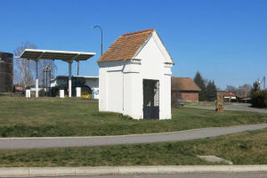 Kleines weißes Gebäude mit einem roten Dach, die erste Tankstelle in Deutschland, neben einer Straße mit Gras, Bäumen und anderen Gegenständen im Vordergrund und Häusern unter einem klaren blauen Himmel im Hintergrund.