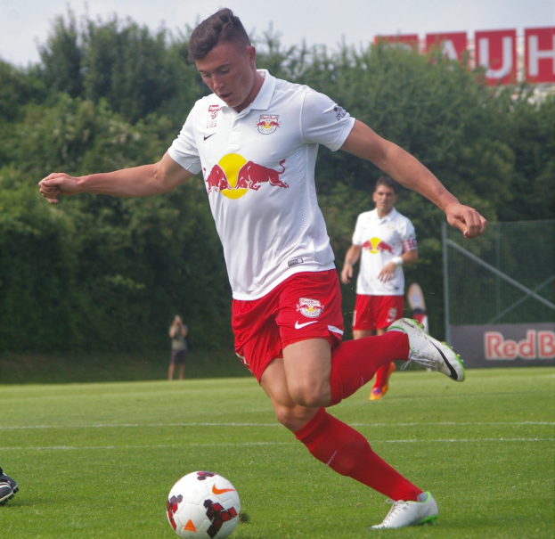 Ein Mann in einem Füßballtrikot schießt einen Ball auf einem grünen Feld mit Bäumen und einem klaren blauen Himmel im Hintergrund, mit einem "RB Leipzig"-Schild im Bild.