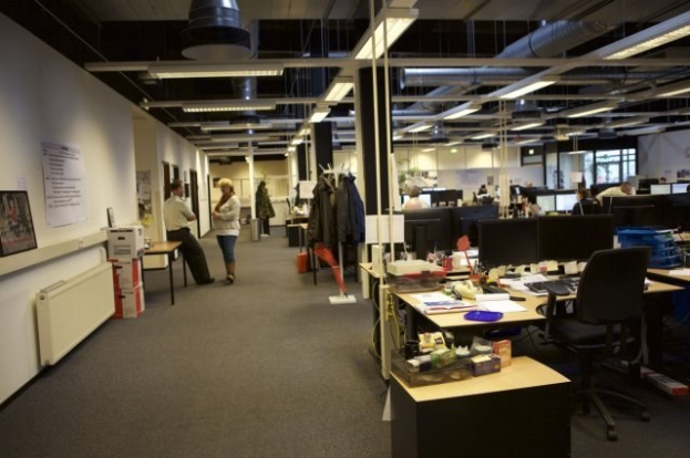 Großes Großraumbüro mit Menschen, die an Schreibtischen arbeiten, Computern und Monitoren, umgeben von Stühlen, Papieren, Büchern und anderen Büromaterialien, mit aufgehängten Kleidungsstücken, Wandtafeln, Deckenlampen und Glasfenstern im Hintergrund.