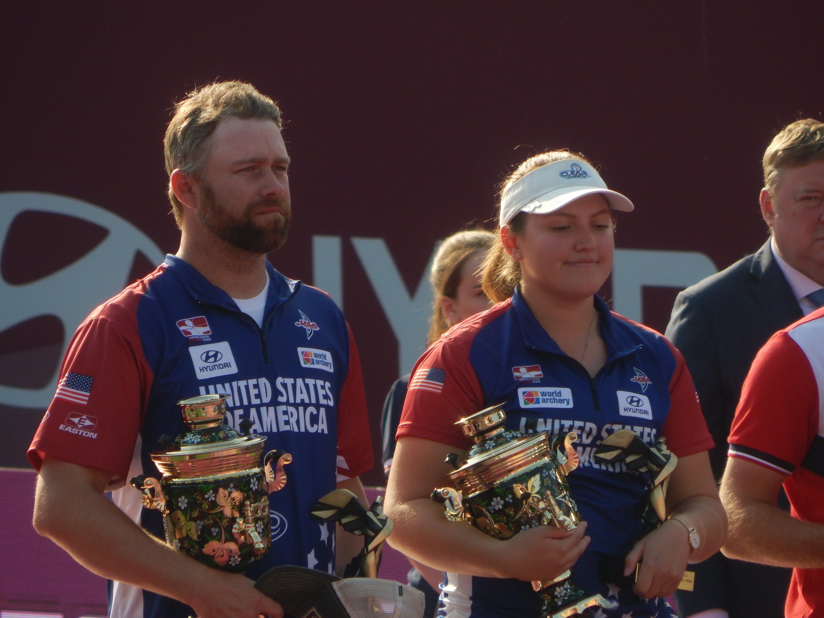 Gruppe von Menschen, die Trophäen halten, mit 'Weltmeisterschaften im Bogensport' -Schilder im Hintergrund.