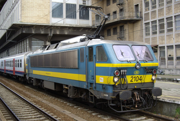 Blauer und gelber Elektrolokomotive, die auf Schienen neben einem Glasgebäude mit einer Plattform und Menschen auf der rechten Seite fährt.