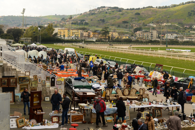 Große Gruppe von Menschen auf einem Flohmarkt mit Tischen, auf denen Gegenstände wie Foto Rahmen und Stühle ausgelegt sind, Fahrzeuge in der Nähe geparkt, Geländer, Treppen, Bäume, Gebäude, Laternenpfähle, Hügel und ein bewölkter Himmel.