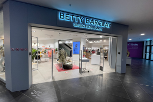 Ein Betty Barclay Selected Brands Geschäft in einem Einkaufszentrum, mit Schaufensterpuppen in der neuesten Mode, Kleidung an Bügeln, Tischen mit verschiedenen Gegenständen, Schildern, einem Topf mit einer Pflanze, Deckenleuchten und einer Tür auf der rechten Seite.