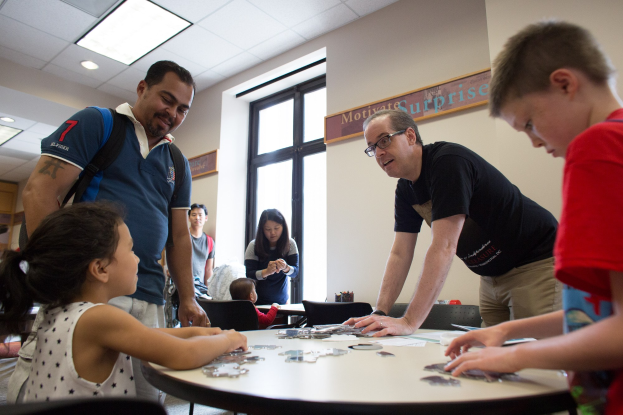 Gruppe von Menschen, die um einen Tisch mit Puzzles, einer Fensterwand mit einer Tafel im Hintergrund und Deckenleuchten sitzen