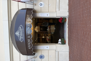 Ein Gebäude mit einer Seite, auf der ein Schild mit der Aufschrift "Boutique & Salon de Monaco" angebracht ist, mit Außenbestuhlung, Blumentöpfen und einem Blick ins gut beleuchtete, luxuriöse Innere.