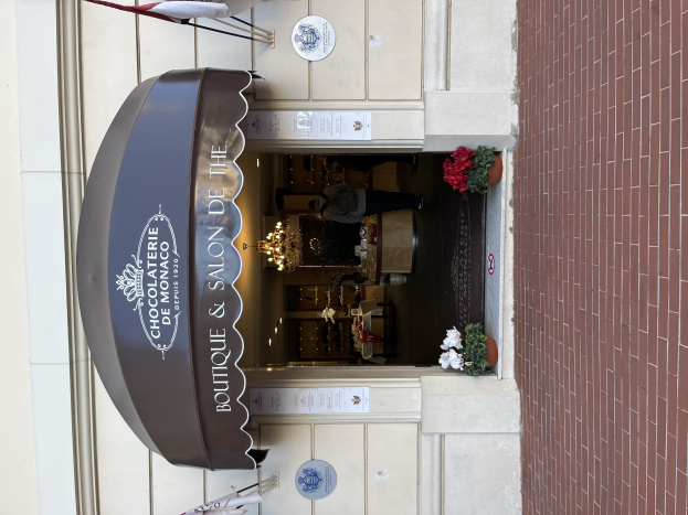 Ein Gebäude mit einer Seite, auf der ein Schild mit der Aufschrift "Boutique & Salon de Monaco" angebracht ist, mit Außenbestuhlung, Blumentöpfen und einem Blick ins gut beleuchtete, luxuriöse Innere.