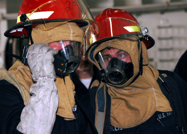 Zwei Feuerwehrleute in Gasmasken und Helmen, einer hält ein Funkgerät in der Hand, stehen vor einer Wand mit freiliegenden Rohren.