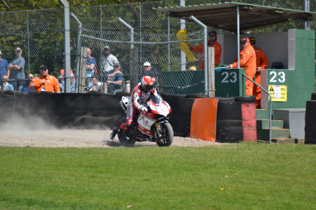 Ein Motorradfahrer in Helm und Handschuhen fährt über ein grasbewachsenes Feld, mit Zuschauern in Mützen und Schutzbrillen, einem eingezäunten Bereich, Treppen mit Geländern und einem nummerierten Schuppen mit Texttafeln im Hintergrund.