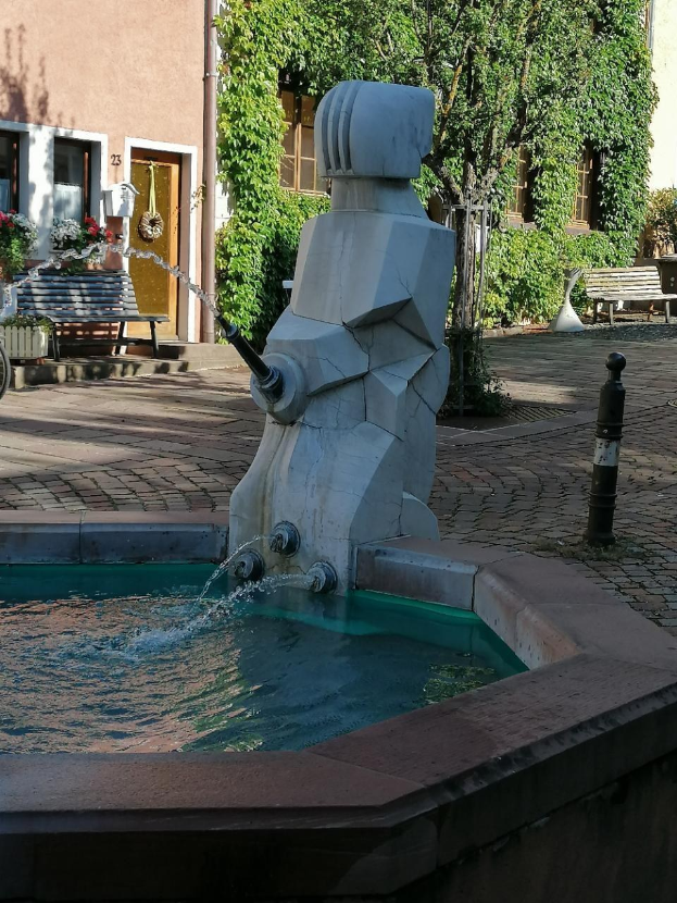 Ein steinerner Brunnen mit fließendem Wasser thront in der Mitte eines Stadtplatzes, umgeben von Gebäuden, Grünflächen, Bänken und Blumenkübeln.