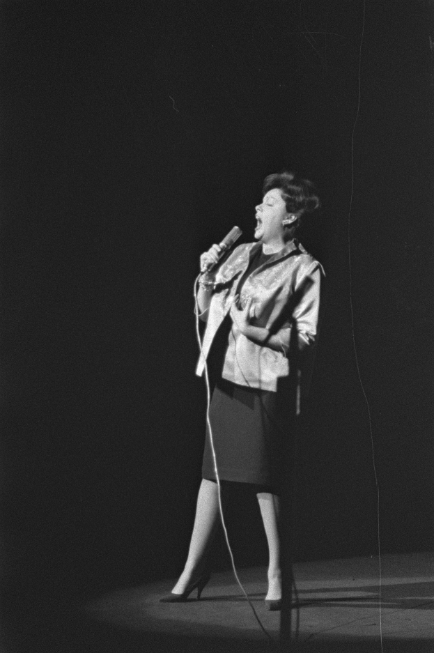 Eine Frau in einem schwarz-weißen Outfit aus den 1950er-Jahren singt fröhlich auf einer von einem Scheinwerfer beleuchteten Bühne und hält ein Mikrofon.