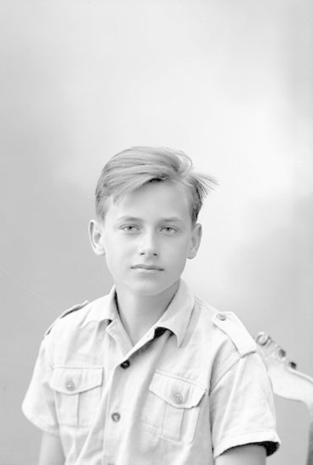 Ein junger Junge in einem Schwarz-Weiß-Foto hält eine Gitarre und trägt ein Hemd mit ernster Miene, kurzen dunklen Haaren und konzentriertem Blick auf das Instrument.