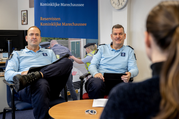 Zwei Polizeibeamte sitzen an einem Tisch mit einer Frau, umgeben von Dokumenten und Büromaterialien, mit einem Banner, einer Uhr und Fotorahmen an der Wand und einem weiteren Tisch mit einem Monitor und Zubehör links daneben.