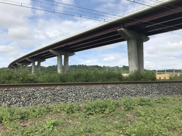 Eine Eisenbahnschiene mit einer Brücke in Renovierung, umgeben von Steinen, Gras, Pflanzen, Bäumen, Strommasten und Drähten, mit einem Gebäude im Hintergrund mit bewölktem Himmel.