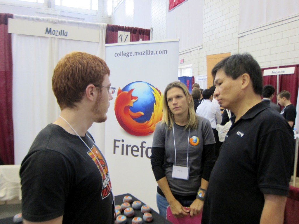 Zwei M├Ąnner und eine Frau stehen vor einem Stuhl und einem Tisch mit runden Gegenst├Ąnden, mit einer wei├čen Bank links, einem wei├čen Banner mit Text dahinter und verschiedenen Bannern, Vorh├Ąngen und einer wei├čen Tafel mit Text im Hintergrund.