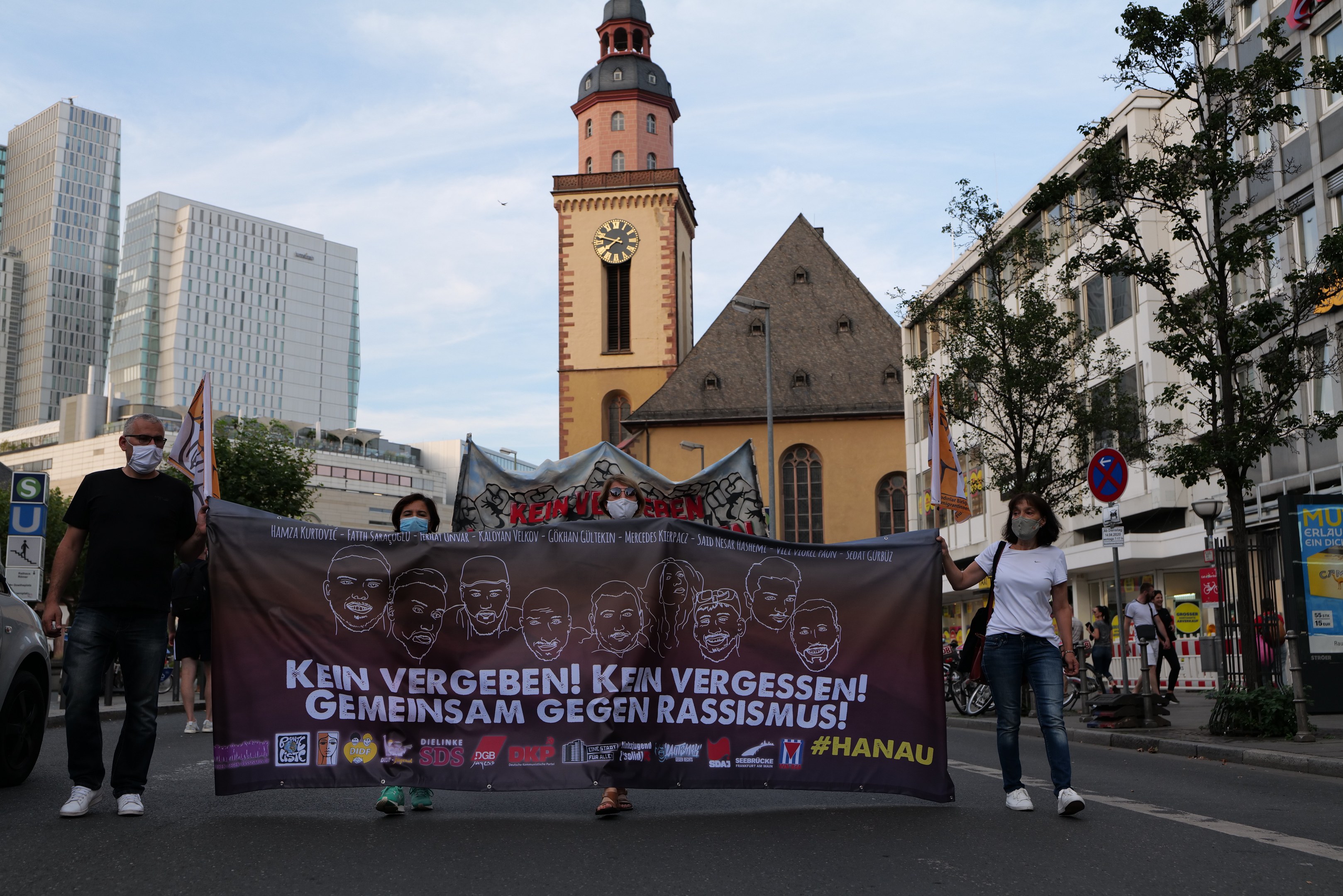 Eine Gruppe von Menschen in Masken, die eine Straße entlanggehen und ein Banner halten, mit einem geparkten Auto auf der linken Seite, Gebäuden und Bäumen im Hintergrund und einem klaren blauen Himmel.