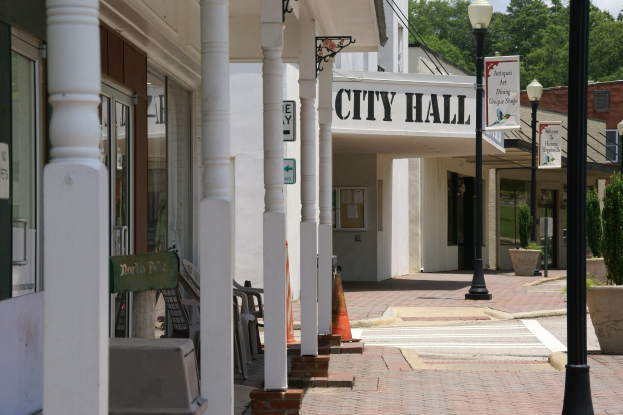 Rathaus mit umliegenden Gebäuden, Straßeninfrastruktur einschließlich Pfosten und Laternen, Namenschilder, fahrende und parkende Fahrzeuge, Verkehrskegel, verstreute Stühle, Topfpflanzen, Bäume und ein bewölkter Himmel.