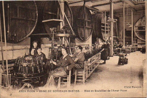 Ein Schwarz-Weiß-Foto von Frauen, die in einer Fabrik arbeiten, mit einigen sitzend und anderen stehend zwischen Maschinen, und Text unten, der 'Amer Picon - Use de Bordeaux-Bastide' liest.