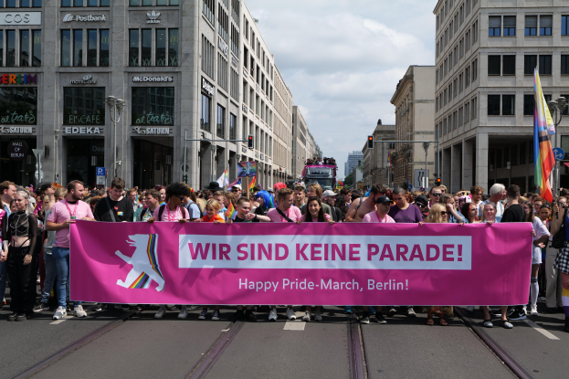 Eine Gruppe von Menschen marschiert auf einer Straße in Berlin, Deutschland, mit einem pinken Banner, auf dem "Happy Pride March" steht, mit Gebäuden, Laternenmasten und Verkehrszeichen an der Straße unter einem bewölkten Himmel.
