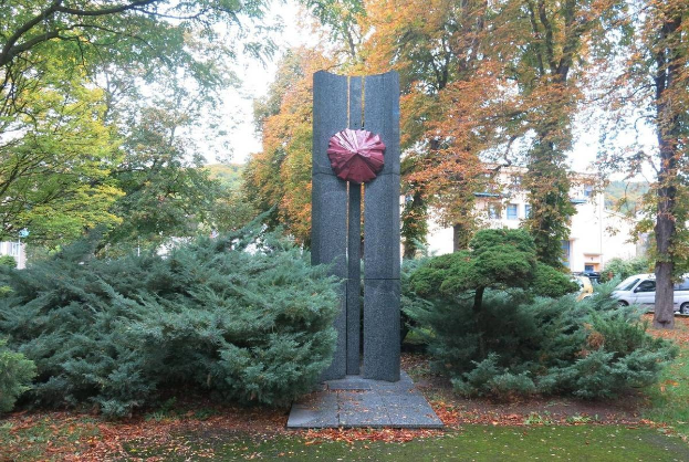 Große Metallskulptur inmitten eines Parks mit Bäumen, Pflanzen und verstreuten Blättern, umgeben von Gebäuden und einem klaren blauen Himmel, Teil der Kunst der Stadt Berlin.
