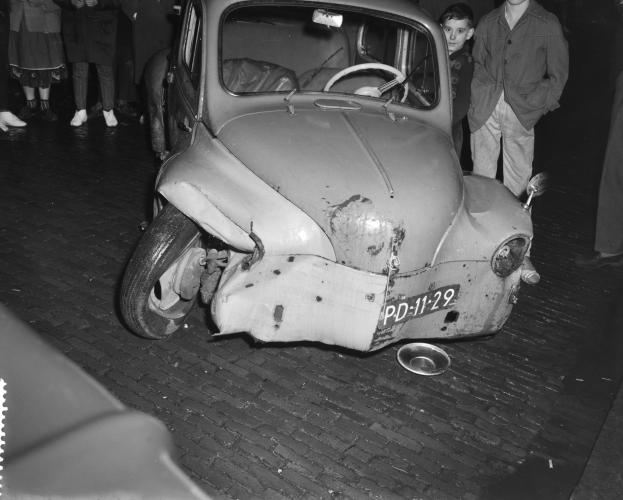 Schwarz-weiß-Foto eines stark beschädigten Autos mit eingedellter Front und zersplitterten Fenstern, umgeben von einer Gruppe von Menschen, die die Szene begutachten.