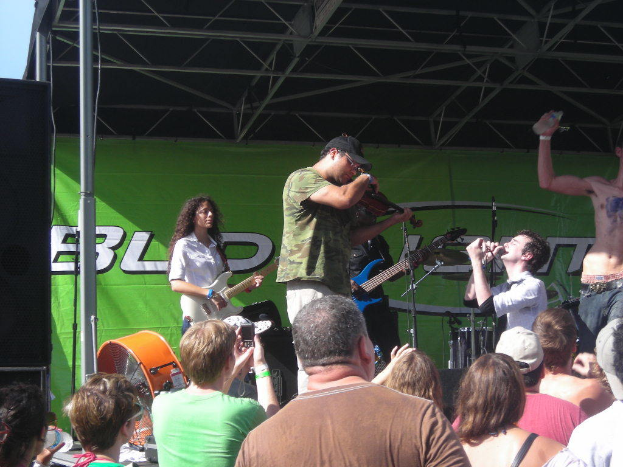 Eine Gruppe von Musikern auf der Bühne, die verschiedene Instrumente spielen, mit einem Publikum davor, einem großen schwarzen Lautsprecher links und einem grünen Banner im Hintergrund.