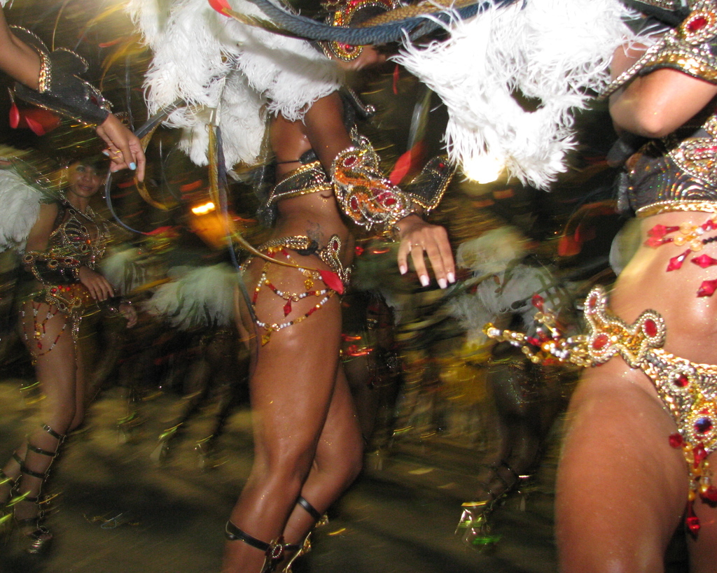 Eine Bank im Vordergrund mit einer verzierten Eisen Säule und Kletterpflanzen dahinter, die eine Frau in einem schwarzen Kleid und ein Gebäude mit Wänden, Fenstern und Türen im Hintergrund zeigt.