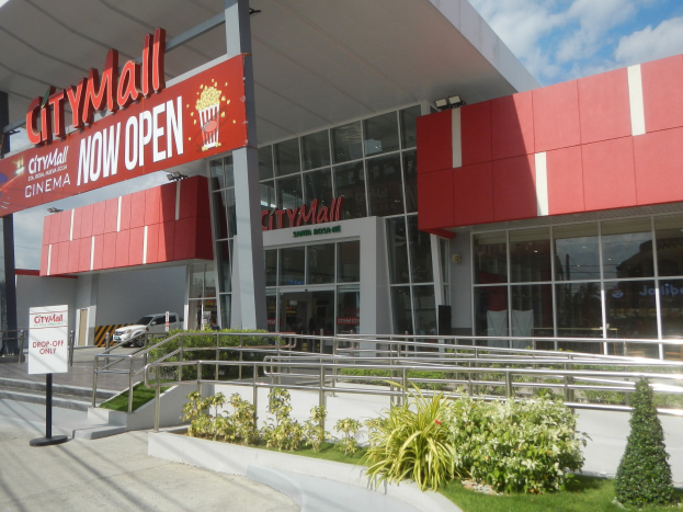 Stadteingangsschild des City Malls mit der Aufschrift "City Mall Now Open" umgeben von Grün, Menschen, einem Auto und einem bewölkten Himmel.