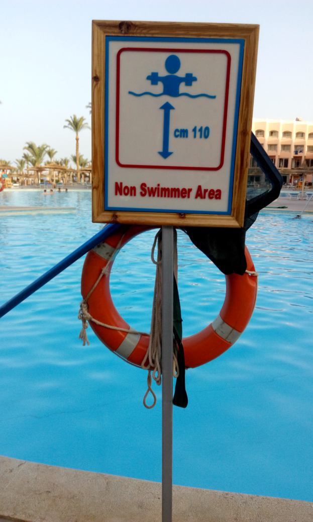 Ein Schwimmbad mit einem "Nicht-Schwimmer-Bereich"-Schild und einem Rettungsring an einem Pfahl, mit Bäumen, Gebäuden und einem klaren blauen Himmel im Hintergrund.