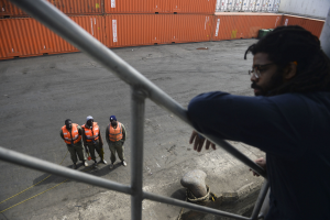 Ein Mann in legerer Kleidung steht auf einem Metallgelönder in der Nähe einer Gruppe von Menschen in orangefarbenen Jacken und Kappen, mit Containern und einer Wand im Hintergrund am Hafen von Los Angeles.