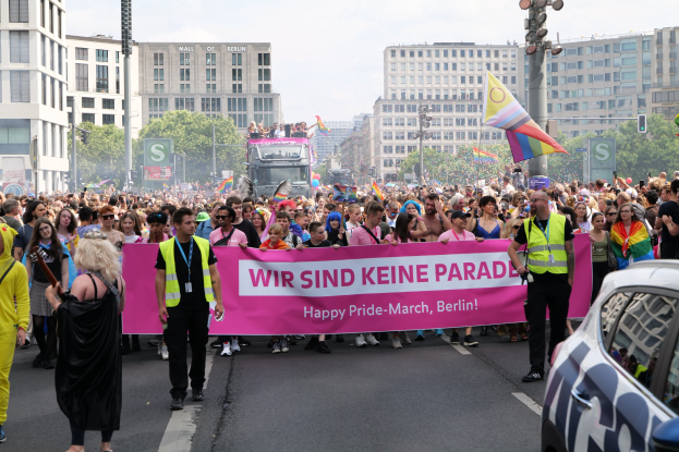Eine große Gruppe von Menschen, die an der 'Happy Pride March Berlin'-Parade teilnimmt und die Straße mit einem Banner öberquert, ein geparktes Auto auf der rechten Seite und im Hintergrund Gebäude, Bäume und einen klaren Himmel.