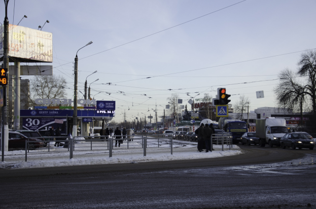 Eine verschneite Straße mit Schwerverkehr, Straßenlaternen, Fahrzeugen, Gelöndern, Polen, Planken, Drähten, Bäumen, Gebäuden und Passanten unter einem bewölktem Himmel.