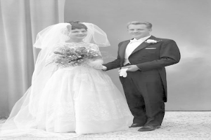 Ein Schwarz-Weiß-Foto eines lachenden Mannes und einer Frau, die an ihrem Hochzeitstag nebeneinanderstehen, wobei die Frau ein weißes Kleid trägt und einen Strauß hält und eine Vorhangwand im Hintergrund zu sehen ist.