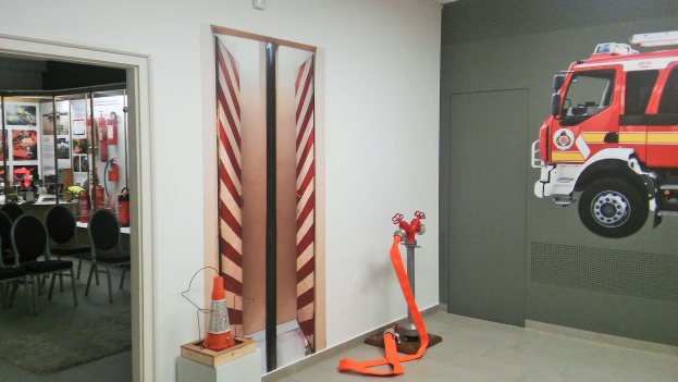 Ein Raum mit einer Wandmalerei eines Feuerwehrautos, einer Tür, einem Verkehrskegel, einem Ständer, einem Teppichboden, Stühlen, Tischen, Feuerlöschern und diversen anderen Gegenständen.