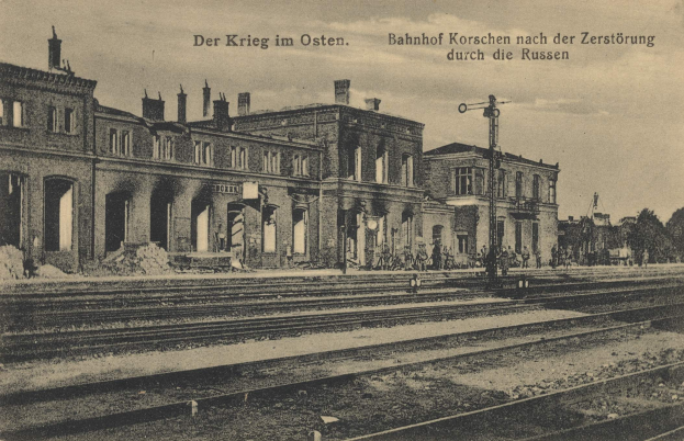 Schwarz-weiß-Foto des Bahnhofs Korschen in Ostern, Deutschland, zeigt Gleise, Gebäude, Pfosten, Menschen, Bäume und Himmel mit Text "Bahnhof Korschen in Ostern, Deutschland" oben und unten.