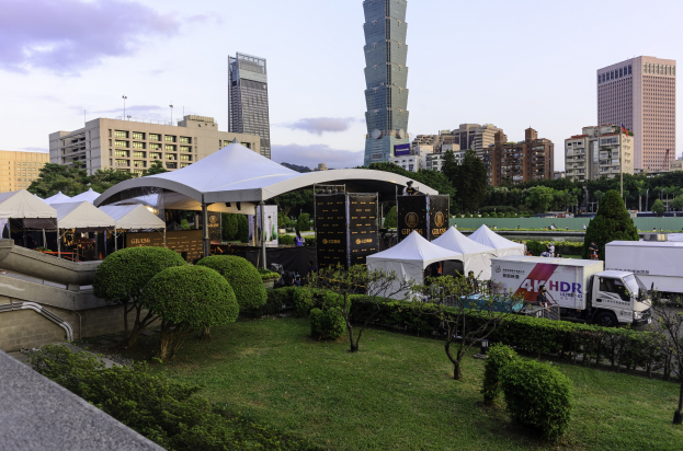 Eine Gruppe von Zelten in einem Park umgeben von Grünanlagen, mit Menschen, Fahrzeugen, Gebäuden und einem klaren Himmel im Hintergrund, wahrscheinlich beim Hong Kong International Film Festival.