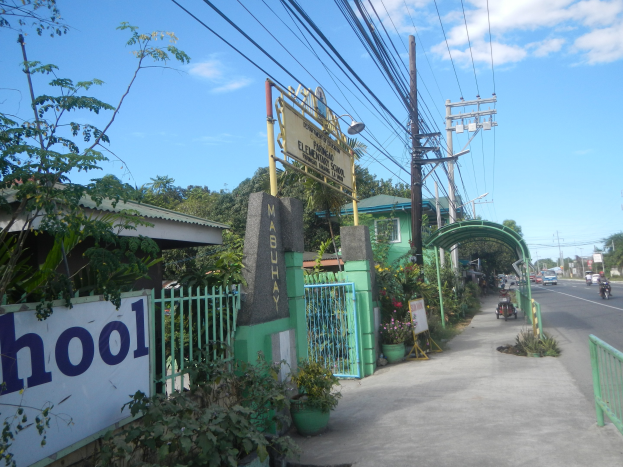 Schuleingang mit einem "Schule"-Schild am Straßenrand, Fahrzeuge, ein Tor, ein Tor, eine Namensschilder, Pflanzen, Bäume, Strommasten mit Drähten und eine bewölkte Himmel.