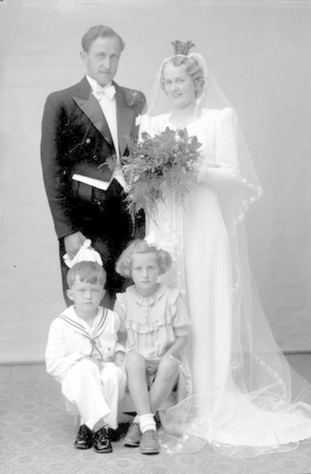 Schwarzes und weißes Foto einer Braut in einem weißen Kleid mit einem Strauß, einem grinsenden Bräutigam neben ihr und zwei Kindern, die vor ihnen auf dem Boden sitzen.