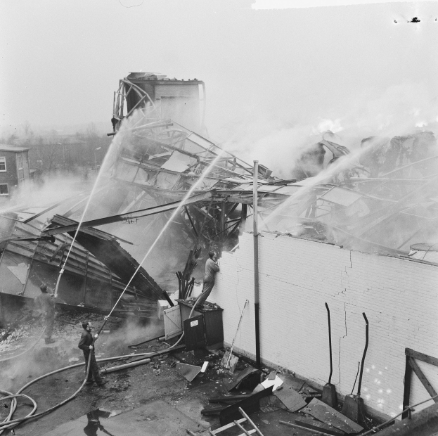 Schwarz-weiß-Foto von Feuerwehrleuten auf einem Dach, die ein Feuer löschen, während Zuschauer unten Equipment halten.